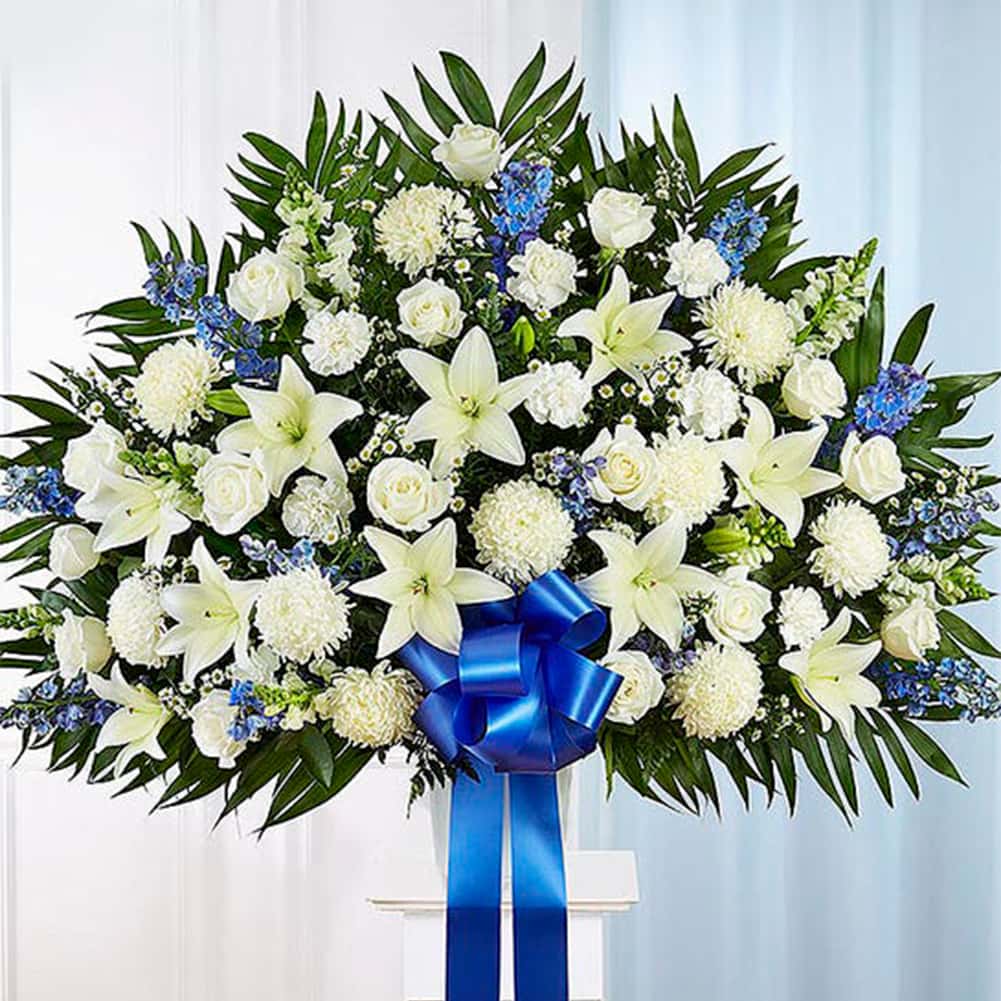 Pedestal Funeral + Basket, Size Extra Large, Fresh Flowers Orlando. Standing basket arrangement with white roses, Asiatic lilies, football mums, snapdragons, carnations and monte casino; blue delphinium; accented with soft, lush greenery and blue bow. Arreglo en canasta para base alta con rosas blancas, lirios asiáticos, crisantemos, boca de dragón, claveles y monte casino; blue delphinium; decoradodo con vegetación en tonos verdes suaves y lazo azul.