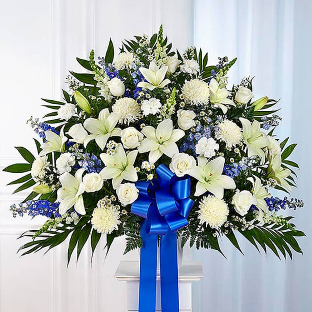 Pedestal Funeral + Basket, Size Large, Fresh Flowers Orlando. Standing basket arrangement with white roses, Asiatic lilies, football mums, snapdragons, carnations and monte casino; blue delphinium; accented with soft, lush greenery and blue bow. Arreglo en canasta para base alta con rosas blancas, lirios asiáticos, crisantemos, boca de dragón, claveles y monte casino; blue delphinium; decoradodo con vegetación en tonos verdes suaves y lazo azul.