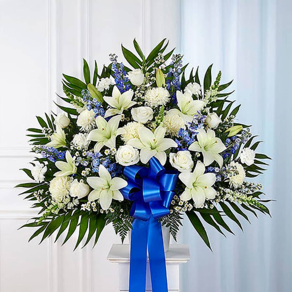 Pedestal Funeral + Basket, Size Medium, Fresh Flowers Orlando. Standing basket arrangement with white roses, Asiatic lilies, football mums, snapdragons, carnations and monte casino; blue delphinium; accented with soft, lush greenery and blue bow. Arreglo en canasta para base alta con rosas blancas, lirios asiáticos, crisantemos, boca de dragón, claveles y monte casino; blue delphinium; decoradodo con vegetación en tonos verdes suaves y lazo azul.