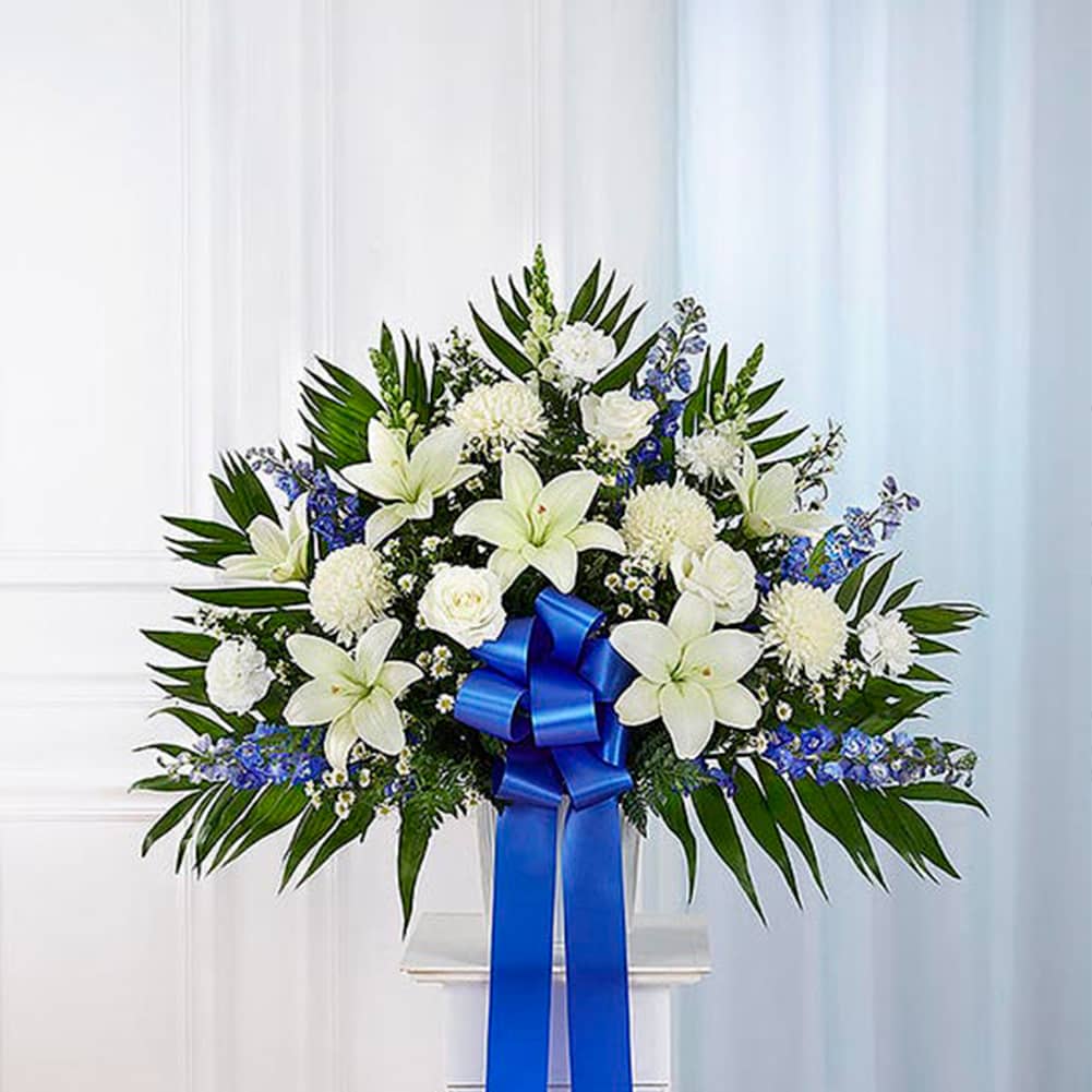 Pedestal Funeral + Basket, Size Small, Fresh Flowers Orlando. Standing basket arrangement with white roses, Asiatic lilies, football mums, snapdragons, carnations and monte casino; blue delphinium; accented with soft, lush greenery and blue bow. Arreglo en canasta para base alta con rosas blancas, lirios asiáticos, crisantemos, boca de dragón, claveles y monte casino; blue delphinium; decoradodo con vegetación en tonos verdes suaves y lazo azul.